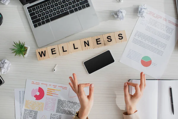 Cropped view of businesswoman making gyan mudra at workplace with documents, laptop, smartphone and alphabet cubes with wellness word — Stock Photo