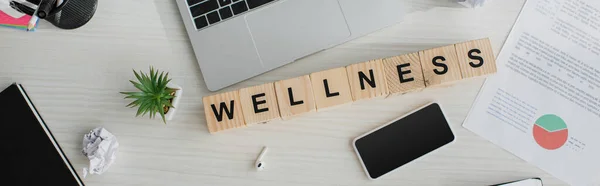 Панорамный снимок рабочего места с наушниками, ноутбуками, смартфонами и кубиками алфавита со словом wellness — стоковое фото