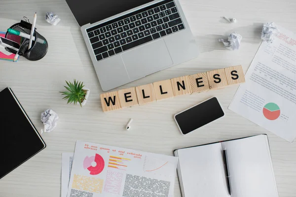 Top view of workplace with business documents, laptop, smartphone and alphabet cubes with wellness word — Stock Photo