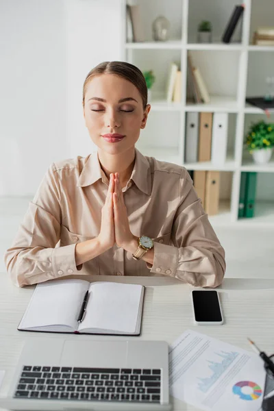 Belle femme d'affaires méditant avec geste namaste sur le lieu de travail avec ordinateur portable, smartphone et bloc-notes — Photo de stock