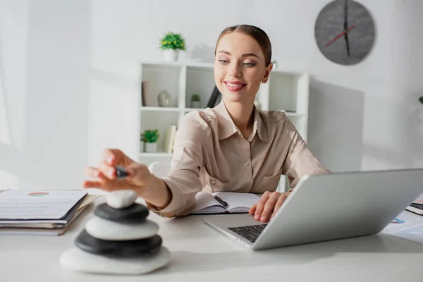 Selective focus of smiling businesswoman working with laptop at workplace with zen stones — Stock Photo