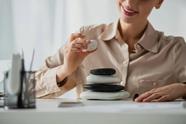 Vista recortada de la mujer de negocios feliz sentado en el lugar de trabajo con piedras zen - foto de stock