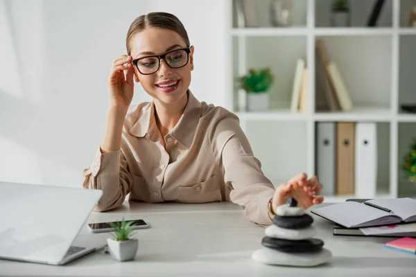 Femme d'affaires souriante assise sur le lieu de travail avec pierres zen, smartphone et ordinateur portable — Photo de stock