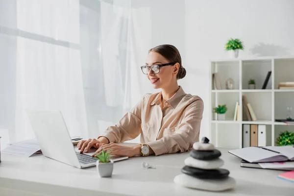 Smiling businesswoman working with laptop at workplace with zen stones — Stock Photo
