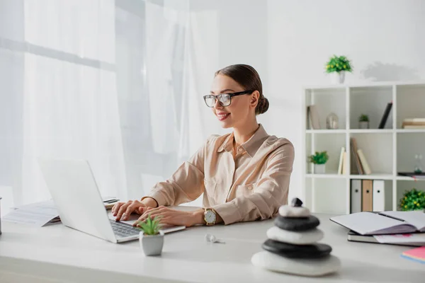 Donna d'affari sorridente che lavora con computer portatile a posto di lavoro con pietre zen — Stock Photo