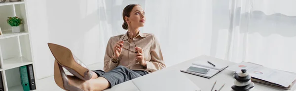 Panoramische Aufnahme einer jungen Geschäftsfrau, die am Arbeitsplatz mit Füßen auf dem Tisch im Büro zögert — Stockfoto