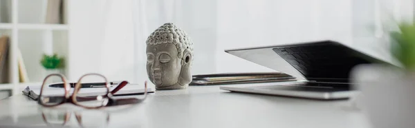 Panoramic shot of Buddha head, eyeglasses and laptop on table in modern office — Stock Photo