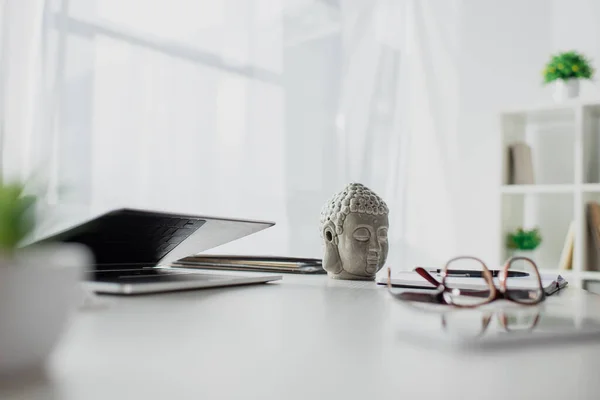 Cabeça de Buda, óculos e laptop na mesa no escritório moderno — Fotografia de Stock