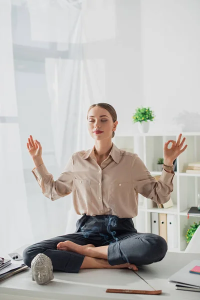 Professionelle Geschäftsfrau meditiert in Lotus-Pose mit Gyan Mudra am Arbeitsplatz mit Buddhakopf und Räucherstäbchen — Stockfoto