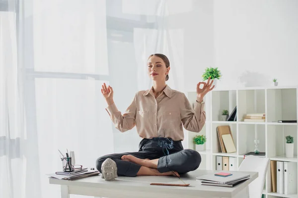 Attractive businesswoman meditating in lotus pose with gyan mudra at workplace with Buddha head and incense stick — Stock Photo