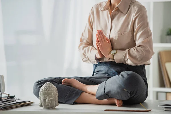 Abgeschnittene Ansicht einer Geschäftsfrau, die in Lotus-Pose mit namaste Geste am Arbeitsplatz mit Buddhakopf und Räucherstäbchen meditiert — Stockfoto