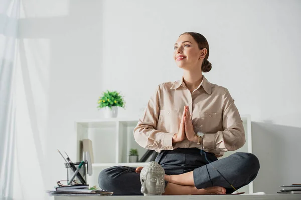 Happy businesswoman meditating in lotus pose with namaste gesture at workplace with Buddha head and incense stick — Stock Photo