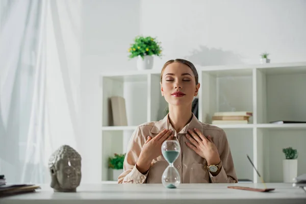 Attractive tired businesswoman sitting at workplace with sand clock and buddha head — Stock Photo