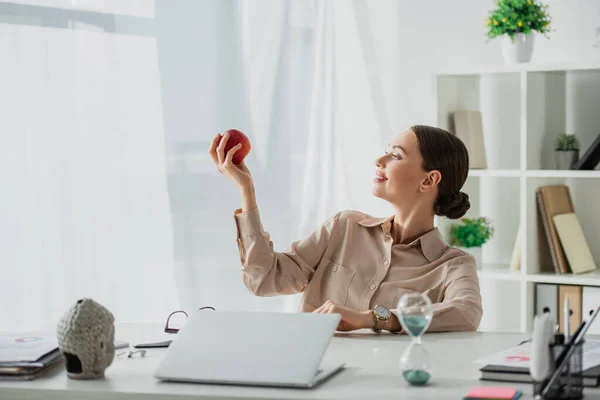 Красива усміхнена бізнес-леді тримає яблуко на робочому місці з головою Будди, ноутбуком і пісочним годинником в офісі — стокове фото