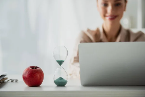 Foyer sélectif de sourire femme d'affaires assis sur le lieu de travail avec pomme, horloge de sable et ordinateur portable dans le bureau — Photo de stock