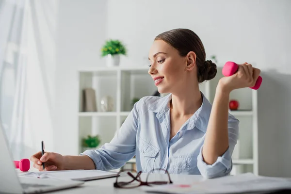Attractive happy businesswoman working while training with dumbbells in office — Stock Photo