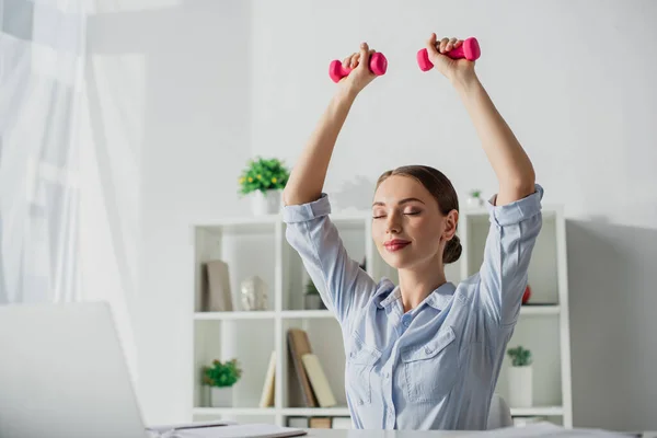 Beautiful happy businesswoman with closed eyes training with dumbbells in office — Stock Photo
