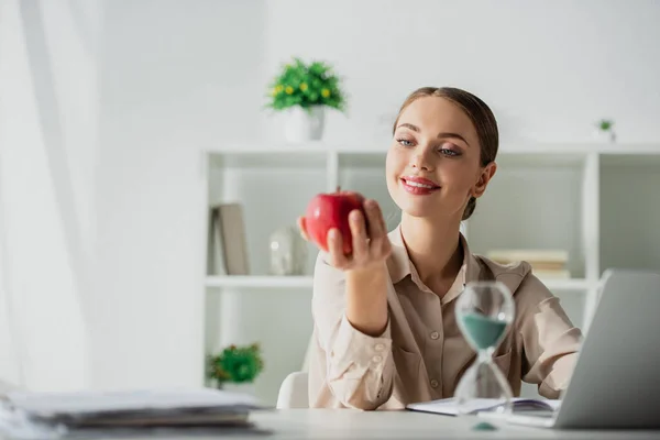 Счастливая деловая женщина держит яблоко на рабочем месте с песчаными часами и ноутбуком в офисе — стоковое фото