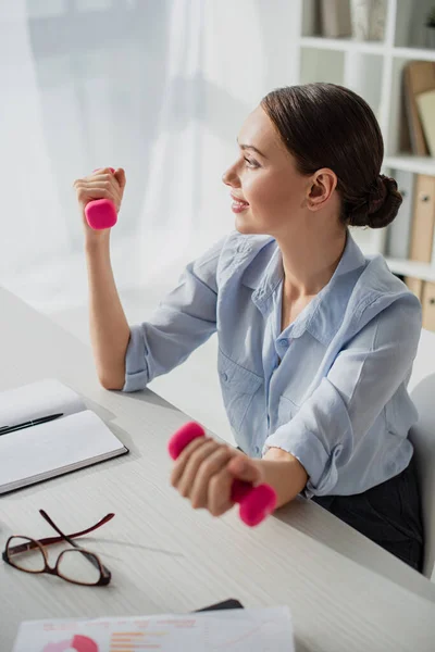 Attractive smiling businesswoman working while training with dumbbells in office — Stock Photo