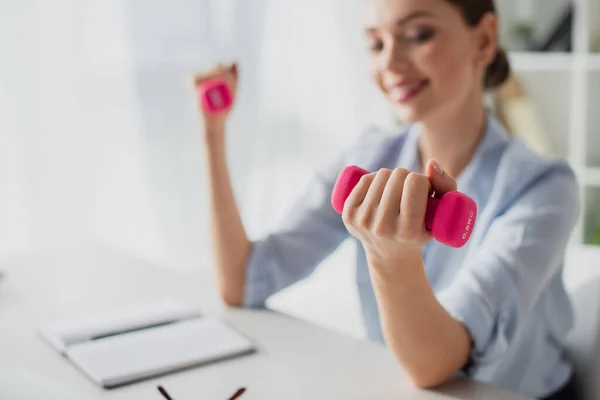 Selective focus of happy businesswoman training with pink dumbbells in office — Stock Photo