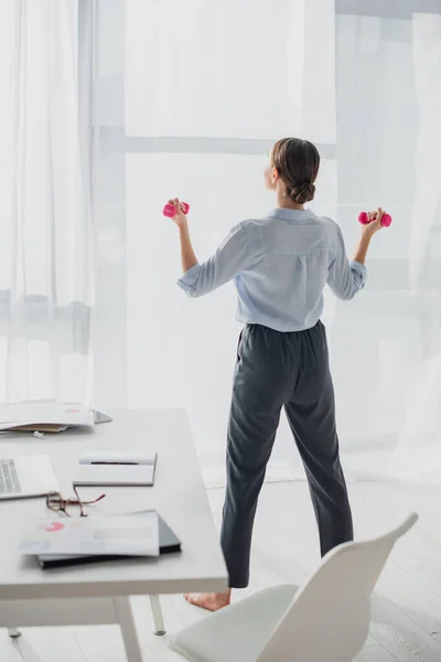 Vista posterior de la joven mujer de negocios de formación con pesas en la oficina con documentos y ordenador portátil - foto de stock