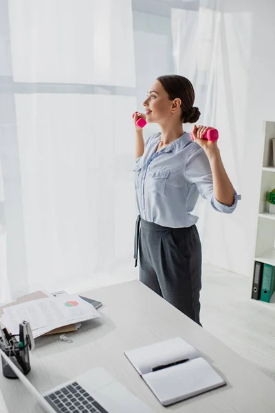 Beautiful businesswoman training with dumbbells in office with documents and laptop — Stock Photo