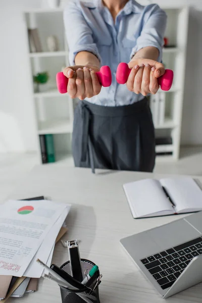 Vista recortada de la formación de mujer de negocios con pesas en el lugar de trabajo con ordenador portátil y papeleo - foto de stock