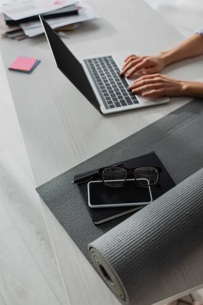 Cropped view of businesswoman working on laptop at workplace with fitness mat, notepad, smartphone and eyeglasses — Stock Photo