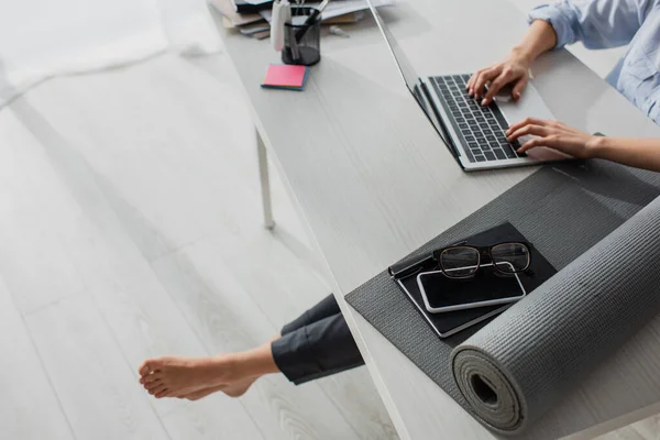 Cropped view of barefoot businesswoman working on laptop at workplace with yoga mat, notepad, smartphone and eyeglasses — Stock Photo