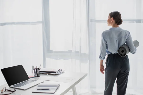 Вид сзади предпринимательницы, держащей коврик для йоги на рабочем месте с ноутбуком и блокнотом — стоковое фото