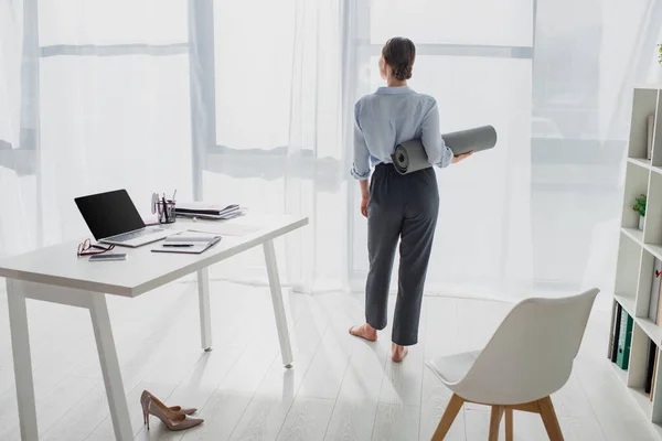 Back view of young businesswoman holding yoga mat at workspace with laptop — Stock Photo