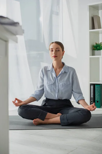 Attractive businesswoman practicing yoga in lotus position with gyan mudra on mat in office — Stock Photo