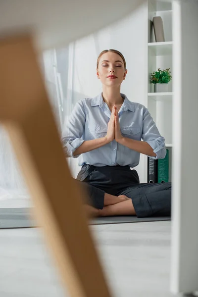 Focalizzazione selettiva della donna d'affari che pratica yoga in posizione di loto con gesto namaste sul tappeto in ufficio — Foto stock