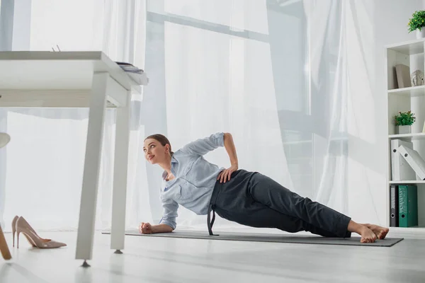 Femme d'affaires souriante pratiquant le yoga en planche latérale sur tapis dans le bureau — Photo de stock