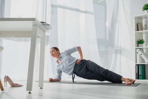 Mujer de negocios sonriente practicando yoga en tablón lateral en la estera en la oficina - foto de stock