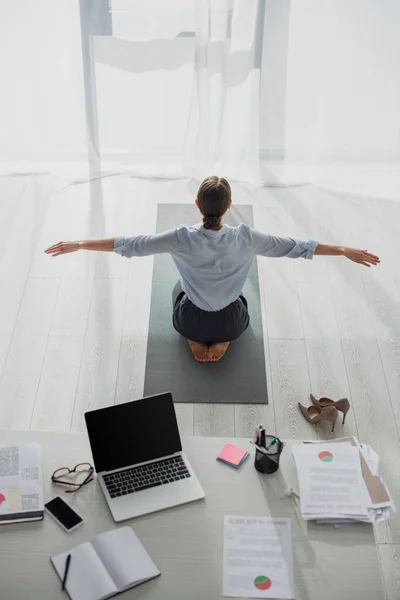 Vista posteriore della donna d'affari che pratica yoga in posizione di loto sul tappeto in ufficio con il computer portatile — Foto stock