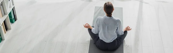 Panoramic shot of businesswoman practicing yoga in lotus position with gyan mudra on mat in office — Stock Photo