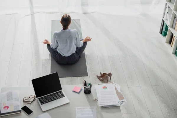 Назад взгляд предпринимательницы практикующей йогу в позе лотоса с гианской мудрой на коврике в офисе с ноутбуком — стоковое фото
