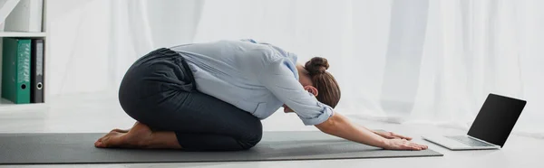 Plan panoramique de jeune femme d'affaires dans Child Pose ayant des cours de yoga en ligne sur ordinateur portable au bureau — Photo de stock