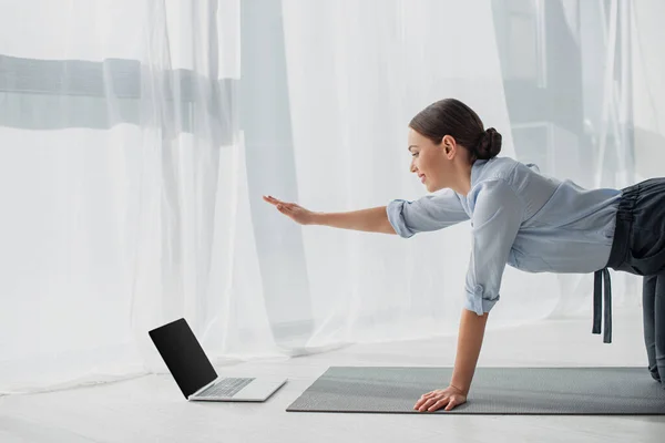 Donna d'affari sorridente che ha lezioni online sul computer portatile e praticare yoga in posa cane uccello sul tappeto in ufficio — Foto stock