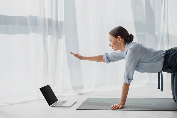 Mujer de negocios sonriente que tiene clases en línea en el ordenador portátil y la práctica de yoga en pájaro perro posan en la estera en la oficina - foto de stock