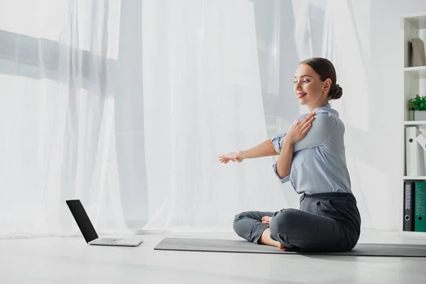 Молода бізнес-леді, що має онлайн-класи на ноутбуці і розтягує руки в позі лотоса на мат в офісі — стокове фото
