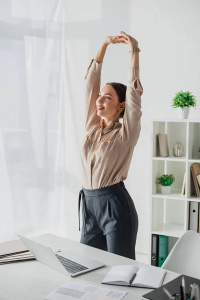 Bela jovem empresária esticando os braços no escritório moderno com laptop — Fotografia de Stock