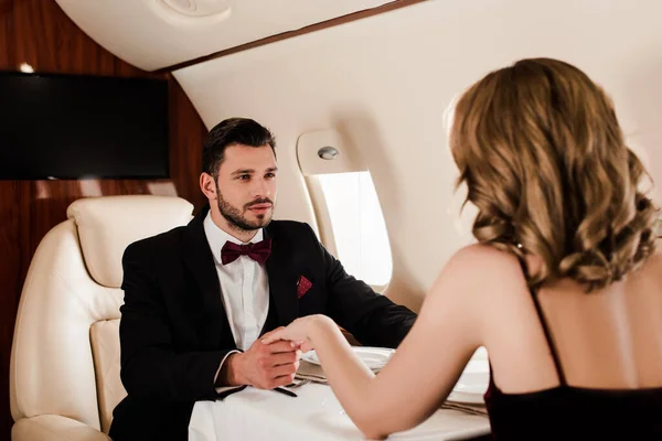 Back view of young woman and elegant man holding hands while sitting at served table in plane — Stock Photo