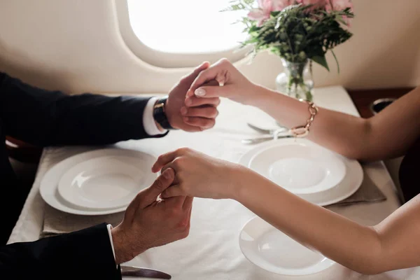 Cropped view of man and woman holding hands while sitting at served table in plane — Stock Photo