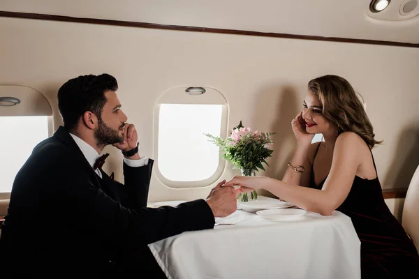 Элегантный мужчина и женщина держатся за руки, сидя за обслуживаемым столом в самолете — стоковое фото