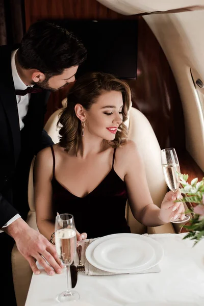 Uomo elegante in piedi vicino ragazza attraente in possesso di bicchiere di champagne in aereo — Foto stock