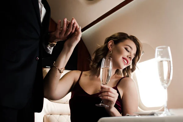 Vista cortada do homem segurando a mão de mulher sorridente, atraente segurando copo de champanhe no avião — Fotografia de Stock