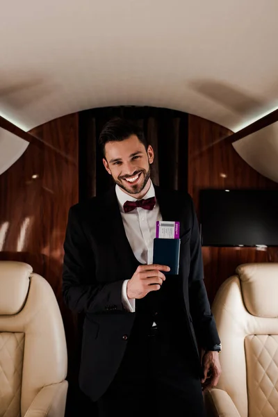 Joven feliz y elegante sonriendo a la cámara mientras sostiene el pasaporte y el billete de avión - foto de stock