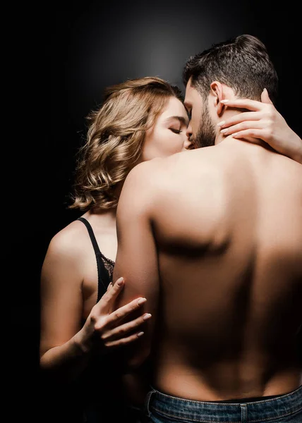 Vista trasera del hombre musculoso sin camisa cerca de chica seductora abrazándolo y besándolo sobre fondo negro - foto de stock